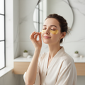 Woman applying gold under-eye patch in bright minimal bathroom